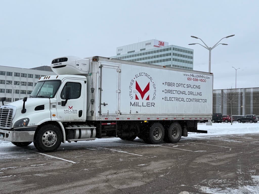A Miller box truck marked for fiber optic splicing, directional drilling, and electrical contracting parked at an urban job site